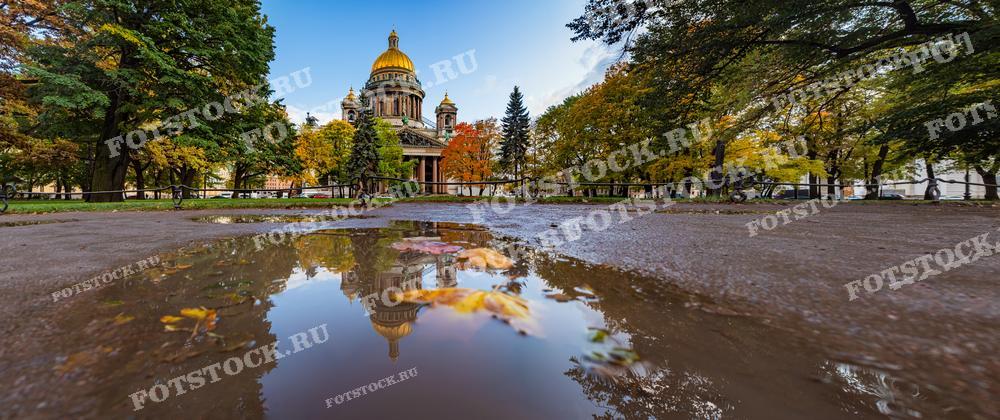 На снимке изображен Исаакиевский собор в Санкт-Петербурге. Фотография сделана в период, когда листья на деревьях окрашены в золотые и красные тона, а отражение собора виднеется в луже на асфальте, что добавляет кадру особенного шарма. Фотографию могут использовать в туристических проспектах осеннего сезона, для статей о путешествиях и как элемент дизайна в интерьере.